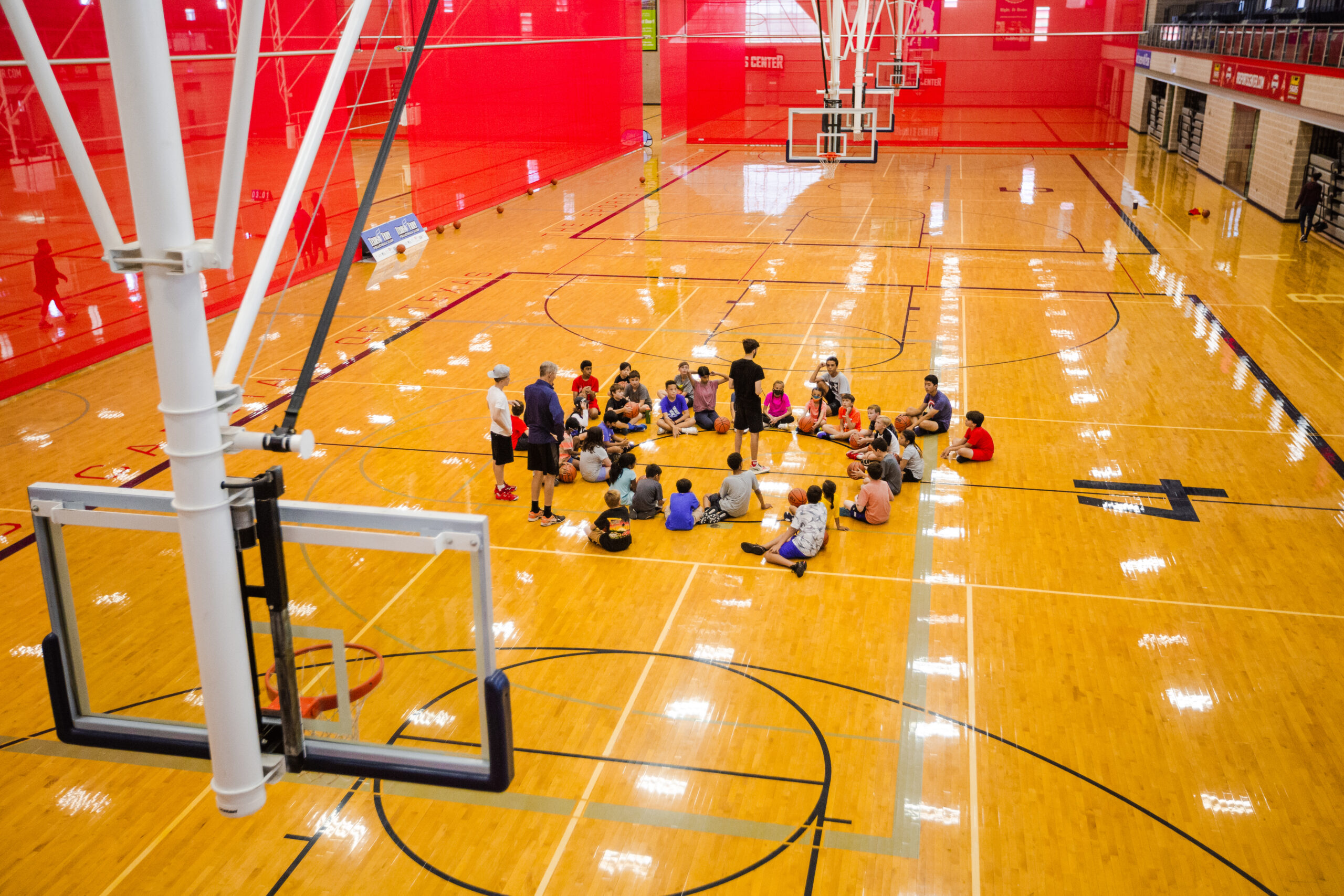 Athletes sitting in a circle on the courts at the Round Rock Sports Center, receiving instruction at a basketball camp